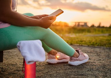 Yakın plan vücut pozitifliği olan sporcu, ağır egzersizden sonra dinleniyor, akıllı telefonuyla mobil uygulamasını kontrol ediyor, spor sahasında terry havlusunun yanında oturuyor, gün batımına karşı suyu olan bir şişenin üzerinde oturuyor.