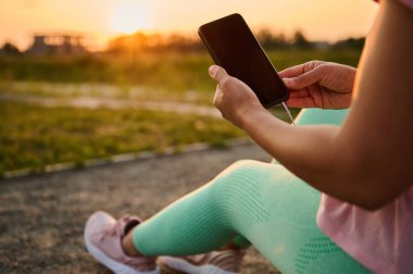Kapalı vücut pozitifliği olan kadın elinde boş siyah ekranlı akıllı telefon, mobil uygulama ve reklam metinleri için reklam alanı olan, spor sahasında oturan ve gün batımında spor yaptıktan sonra dinlenen