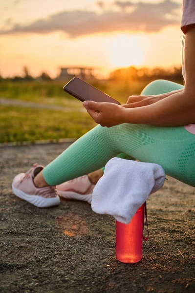 Yakın plan atlet elinde mobil uygulama ve reklam metni için reklam alanı olan boş siyah ekranlı akıllı telefonu tutuyor, spor sahasında oturuyor ve gün batımında antrenmandan sonra dinleniyor