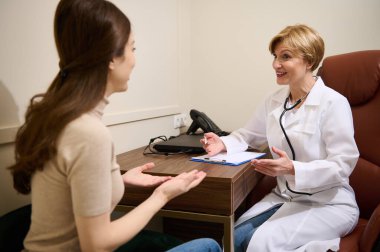Deneyimli kadın pratisyen hekim genç bir kadın hastaya danışıyor, tıbbi form dolduruyor ve klinikteki randevuda ilaç yazıyor..