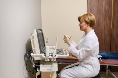 Sevimli kadın radyoloji doktoru bir elinde ultrason sondası tutuyor ve modern ultrason makinesindeki hastayı incelerken monitör ekranına bakıyor..