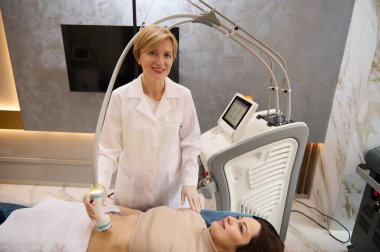 Donanım kozmetolojisi. Kendine güvenen kadın kozmetik uzmanı, estetisyen, güzellik salonunda genç ve güzel bir bayana RF kaldırma prosedürü uyguluyor.