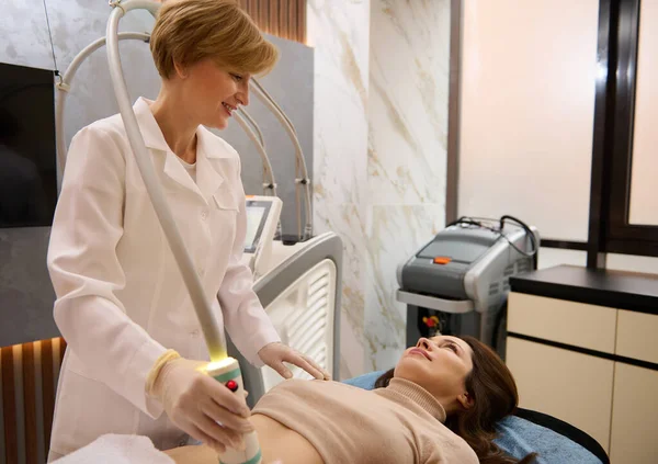 Hoş bir güzellik uzmanı güzel bir kadının karın kasları üzerinde radyo frekansı sensörünü hareket ettiriyor. Mikrostimülasyon, anti-selülit tedavisi ve sağlık kaplıcalarında kilo kaybı için.