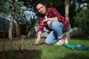 Middle-aged Hispanic pretty woman gardener smiles looking at camera while loosening the black soil with a gardening rake. Horticulture, hobby and leisure activity