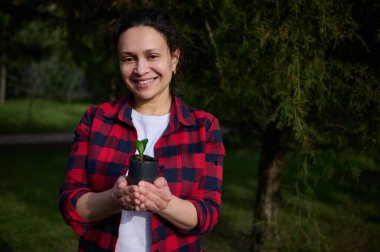 Bir kır evinin bahar bahçesinde duran ve elinde siyah toprakta filizlenen bir çaydanlık tutan gülümseyen kadın bahçıvan. Çiftçilik ve tarım hobisi, bahçıvanlık, bahçıvanlık