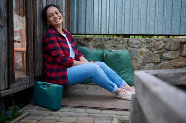 Büyüleyici orta yaşlı bahçıvan, iki büyük gübre torbasının arka planına karşı elinde bahçe aletleri olan bir valizin yanındaki ahşap seranın girişinde oturuyor.