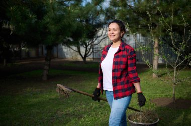 Beyaz tişörtlü, ekose gömlekli, iş eldivenli, elinde yaba ve çim biçme kovası olan çalışkan kadın bahçıvan.
