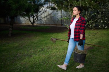 Bir kadın bahçıvan bahçede çalışıyor, elinde çimleri biçilmiş bir kova ve bir yaba ile bölgeyi temizliyor.