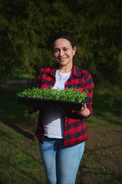 Zevkli, çok ırklı, orta yaşlı, kareli gömlekli, beyaz tişörtlü, kot pantolonlu, elinde fidelerle kaset tutan orta yaşlı bir kadının portresi.
