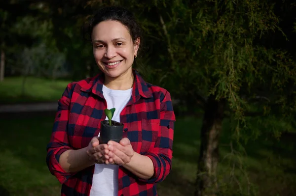 Bir kır evinin bahar bahçesinde duran ve elinde siyah toprakta filizlenen bir çaydanlık tutan gülümseyen kadın bahçıvan. Çiftçilik ve tarım hobisi, bahçıvanlık, bahçıvanlık