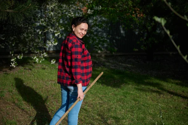 Orta yaşlı, etnik çeşitlilikte bir kadın, hoş bir ev hanımı ve bahçıvan. Bahçede çalışırken kameraya bakıp gülümsüyor.