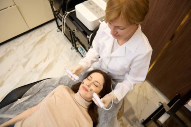 Güzellik terapistinin güzellik salonunda orta yaşlı çekici bir kadına mikro akım tedavisi uygulaması. Donanım kozmetolojisi, profesyonel yaşlanmayı önleme ve cilt bakımı konsepti