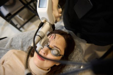 Beautician aesthetician using laser handle on middle aged woman face while making laser skin resurfacing in aesthetic medicine