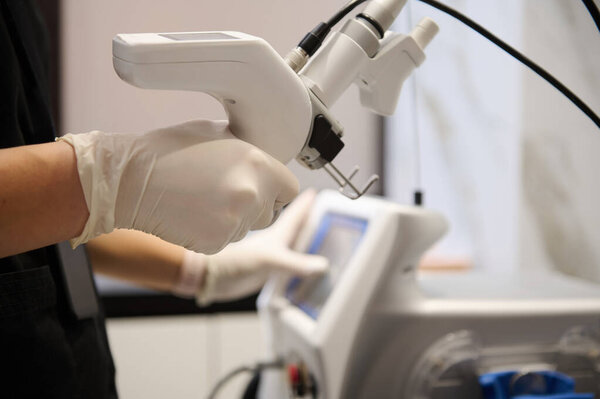 Soft focus on the hands of an aesthetician in a black uniform holding a laser resurfacing machine and pointing with finger at the monitor screen and adjusting programs on a hardware beauty apparatus