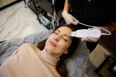 Çekici, orta yaşlı, güzel, koyu saçlı, beyaz bir kadının, sağlık merkezindeki ultrason dişlerini soyarken çekilmiş bir fotoğrafı. Cilt bakımı, profesyonel güzellik tedavi konsepti