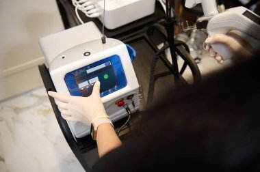 Siyah üniformalı bir estetisyenin elinde lazer yüzey makinesiyle ekranı gösteren ve programları donanımlı bir güzellik aparatına yerleştiren ellerinin tepe noktası.