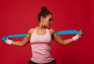Afrikalı Amerikalı kararlı orta yaşlı, kaslı kadın dar pembe bluzlu ve terry bileklikli. Kollar üzerinde esneme egzersizleri yapıyor. Kırmızı arka planda elastik fitness bandı kullanıyor.