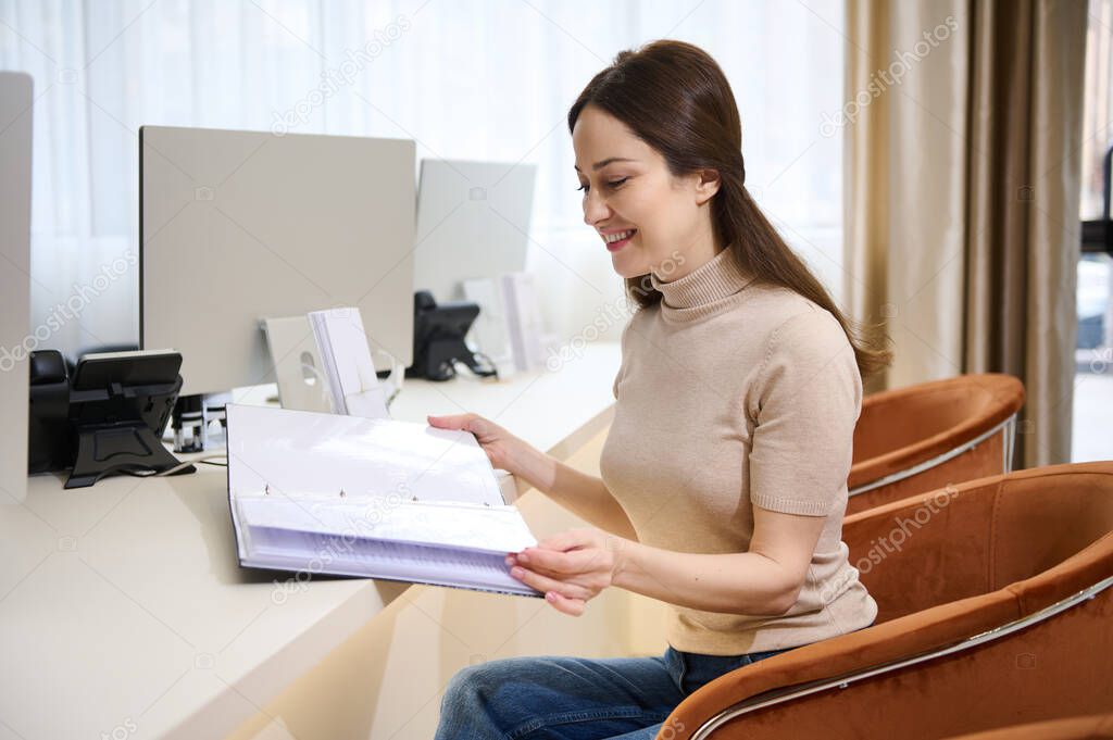 Attractive young dark-haired Caucasian woman in casual clothes browsing a catalog of services while sitting in a comfortable chair at a desk at the reception of a medical clinic