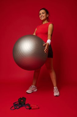 Spor giyim sektöründe çalışan güzel bir kadının tam boy portresi. Reklam metni için fotokopi alanı olan kırmızı renkli arka planda uygun bir topla egzersiz yapıyor. Spor, aktif yaşam tarzı, fitness konsepti