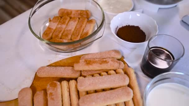 キッチンテーブルに置かれたクラシックなイタリアのティラミスデザートを作るための食材のウォークスルービデオ ティーフィンガービスケットクリームチーズエスプレッソコーヒーミルククリームココアパウダー — ストック動画