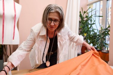 Tecrübeli bir terzi bir tekstil seriyor. Hoş, olgun Avrupalı kadın moda tasarımcısı yeni elbiseler üretmek için yeni kumaşlar seriyor. Terzilik. Moda tasarım atölyesinde kıyafet yapmak