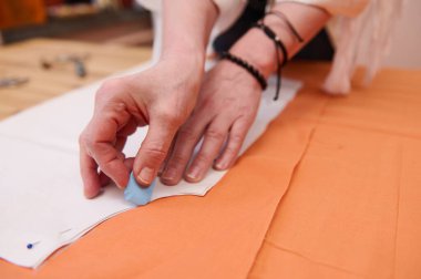 Bir moda stüdyosunda turuncu renkli bir kumaşa tebeşir ya da sabunla dikiş deseni çizerken bir terzinin elinin ayrıntıları. Moda atölyesi, terzilik, el yapımı kıyafet konsepti..