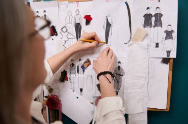 Moda tasarımcısının eline kalem tutarken ve duvara çizimler ve esanslarla tutturulmuş giysiler çizerken dikkat edin. Yaratıcı mesleki meslek kavramı