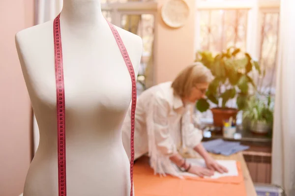 Soft focus on a mannequin and meter in fashion clothing design atelier on the blurred background of a stylish elderly female tailor working with fabric and sewing pattern while creating new garment