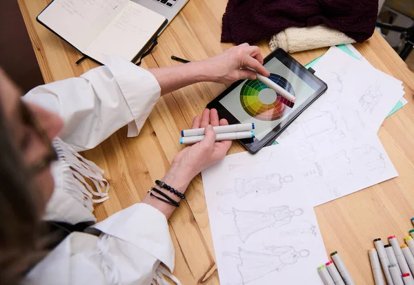 Overhead view. Mature European confident woman, senior fashion designer choosing color palette. Matches color markers to the color wheel while coloring clothes sketches of new collection