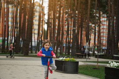 Güneşli bir sonbahar ya da baharın başında şehir parkında scooter kullanan sevimli beyaz bir kız. Sağlıklı ve aktif yaşam tarzı, boş zaman aktivitesi, mutlu çocukluk konsepti