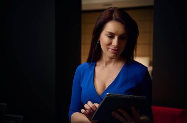 Yakın plan portre, zarif orta yaşlı, güzel, koyu renk saçlı, mavi elbiseli Avrupalı bir kadın. Tasarımcı, iç tasarım stüdyosunda dijital tablet üzerinde çalışıyor.