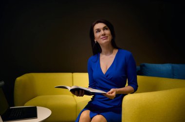 Sarı koltukta oturan ve elinde katalog tutan, sergi salonundaki yeni döşenmiş mobilyaları seçerken rüya gibi bakışlar atan hoş, çarpıcı, çekici, zarif, beyaz tenli, esmer kadın.