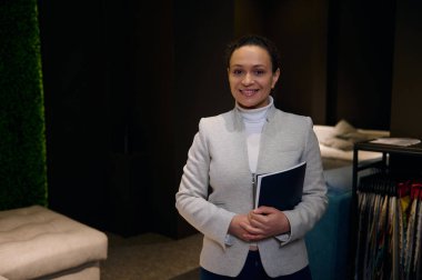 Afrikalı güzel bir kadın, satış müdürü, iç mimar mobilya tasarım galerisinde döşeme kumaşı örnekleriyle tezgahın yanında duruyor, bir katalog tutuyor ve kameraya güvenle bakıyor.
