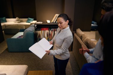 Çok ırklı orta yaşlı kadın satış asistanı gündelik giysiler içinde mobilya koleksiyonunu mobilya alışveriş merkezindeki birkaç müşteriye katalogda sunuyor.
