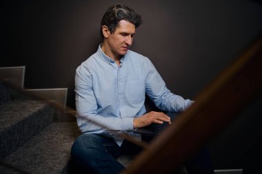 Kendinden emin deneyimli Kafkas girişimci, resmi giyimli iş adamı ev içi merdivenlerde dizüstü bilgisayarda çalışıyor. Uzak meslek, çevrimiçi iş ve proje kavramı.