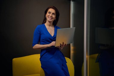 Göz kamaştırıcı şık giyimli Avrupalı bir kadın dizüstü bilgisayarla uzaktan çalışıyor, sarı yeni sanat sandalyesine yaslanıyor ve modern ofise hayali bir şekilde gülümsüyor. İş kadını, başarı, online