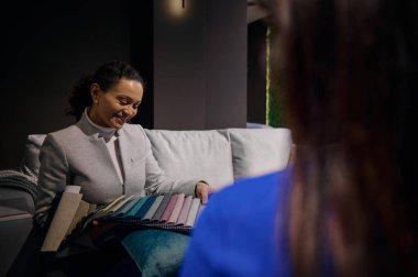 Çok ırklı kadın satış müdürü, müşterilerle çalışan iç mimar, kanepe döşemesi ve restorasyonu için çeşitli doku ve kalitede renkli kumaş örnekleri gösteriyor.