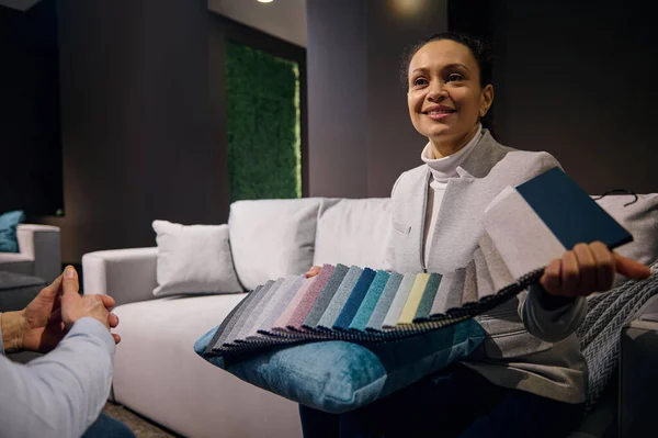 Confident pleasant African American woman, interior designer sales manager in furniture showroom holding colorful fabric samples for soft furniture upholstery and renovation. Home design project