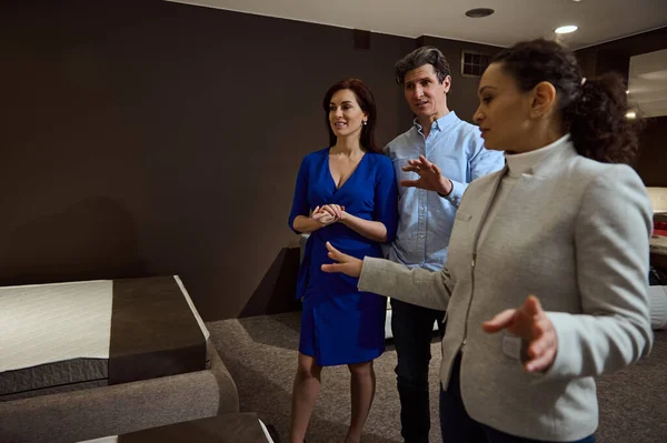 In blurred foreground experienced confident sales consultant, clerk, saleswoman shows various bedding items to married couple shopping for a sleeping bed and orthopedic mattress in furniture store