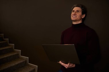 Koyu kahverengi duvar arka planında dizüstü bilgisayarı olan yakışıklı, olgun, beyaz bir adam. İş adamı, girişimci, metin yazarı, gazeteci, serbest yazar. Uzak ve çevrimiçi çalışma kavramı