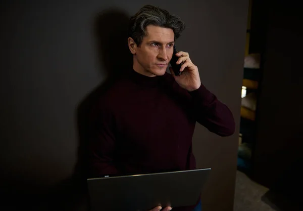 Handsome successful multitasking European businessman, entrepreneur, copywriter talks on mobile phone and works online on laptop standing against a dark wall lit with dim light in a modern office hall