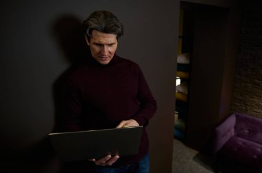 Çok yönlü başarılı beyaz iş adamı, girişimci, metin yazarı modern ofis içinde loş ışıkla aydınlatılmış karanlık bir duvara karşı duran bir dizüstü bilgisayarda çalışıyor.