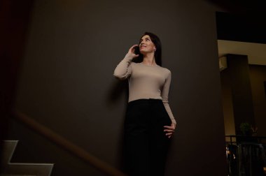 Oldukça koyu saçlı, beyaz bir kadın, iş kadını, günlük giysiler içinde tasarımcı cep telefonuyla konuşuyor, modern bir iç mimari ofisinin koridorunda dedikodu yapıyor. Alt görünüm