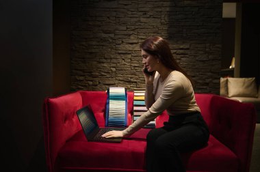 Dizüstü bilgisayar üzerinde çalışırken cep telefonuyla konuşan, yeni ev tasarımı projesi planlayan, döşeme kumaşı örnekleriyle döşenmiş bir kanepede oturan deneyimli kadın iç mimar.