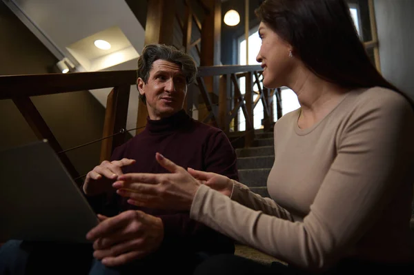 They are great team together. Friendly colleagues, business partners planning new projects sitting close to each other on stairs in a modern office lit with dim light