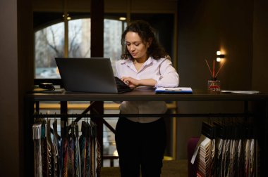 Hoş bayan profesyonel dekoratör, ev tasarımcısı rahat yaratıcı stüdyosunda dizüstü bilgisayar üzerinde çalışıyor, yeni projesi için kumaş örnekleriyle bir standın yanında duruyor.