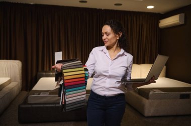 Yaratıcı kadın profesyonel dekoratör yeni ev tasarımı projesinde çalışıyor, dizüstü bilgisayarını elinde tutuyor ve döşeme kumaşlarını seçiyor, mobilya mağazasındaki döşemelere karşı duruyor.