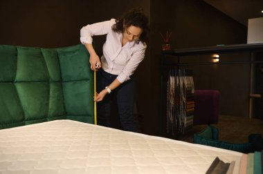 Sıradan giyinen profesyonel tasarımcı ölçüm bandı kullanarak mobilya tasarım stüdyosundaki yumuşak bir yatağın boyutlarını ölçüyor. Ev dekorasyonu, mobilya restorasyonu ve ev tasarımı konsepti