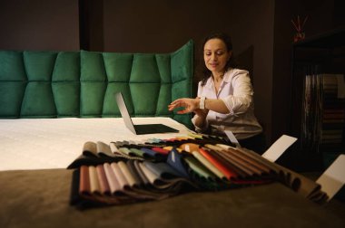 Kadın iç mimar, dekoratör, satış müdürü. Ev tasarımı stüdyosundaki renkli kumaş örneklerinin yanında ortopedik yatak üzerinde çalışırken saatini kontrol ediyor.