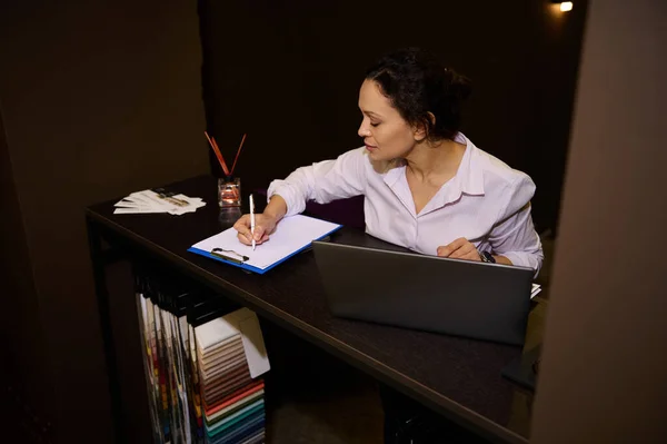 Inspired female decorator interior designer planning home design project, drawing sketch while working laptop in a furniture design creative studio near stand with upholstery fabric samples